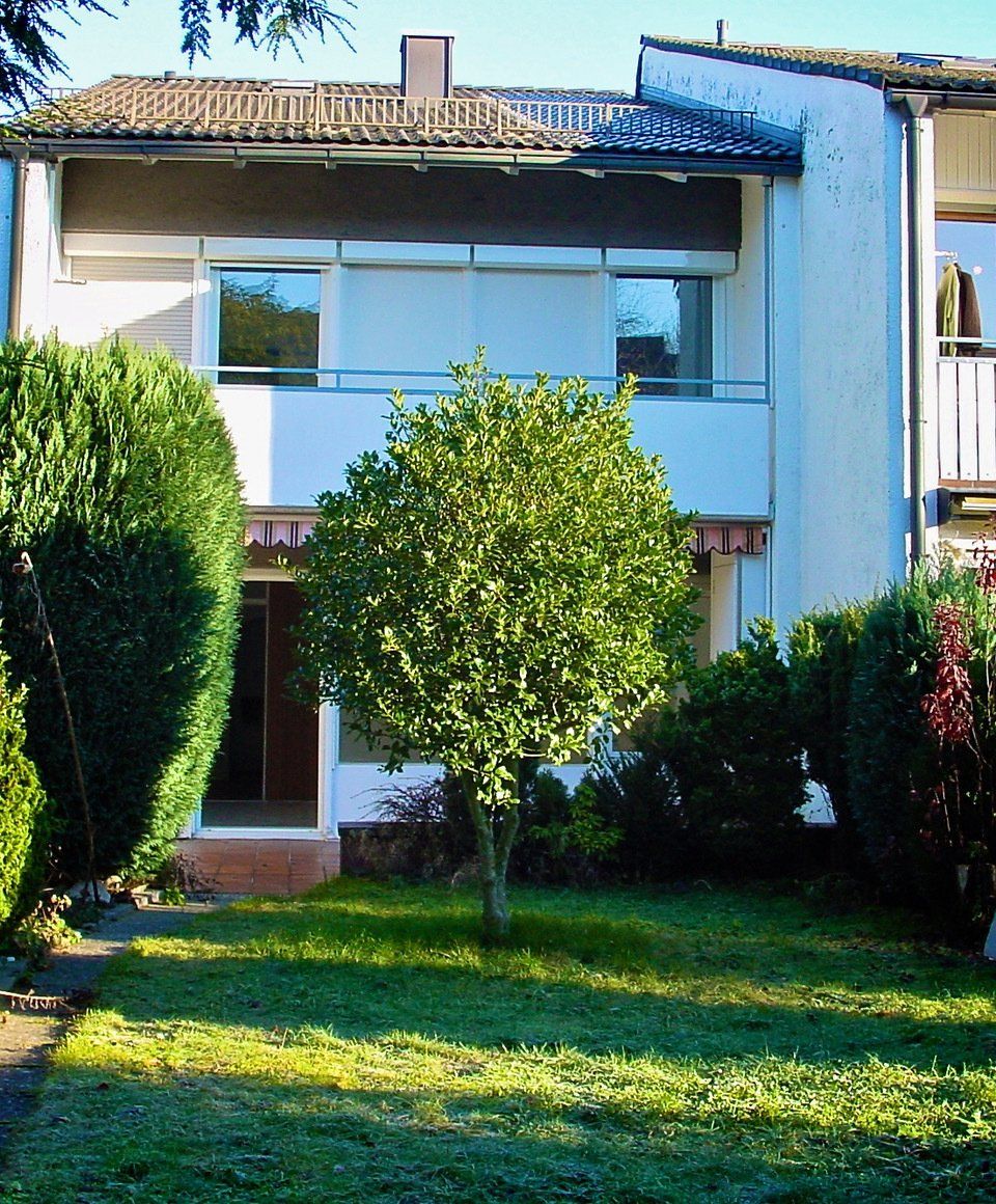 Reihenhaus mit Balkon, kleinem Baum und grünem Rasen.