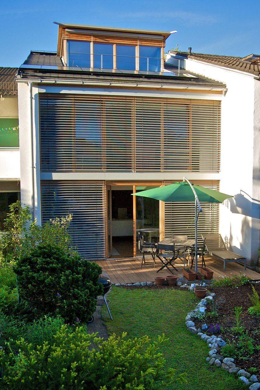 Zweistöckiges Haus mit Holzlamellen vor den Fenstern. Hinterhof mit Terrasse, Tisch, Sonnenschirm und Garten.