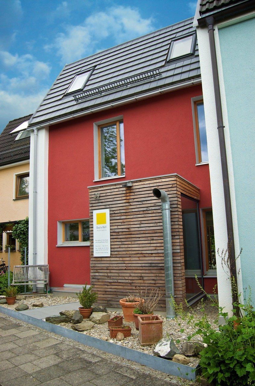 Rotes zweistöckiges Haus mit grauem Dach, hölzernem Seiteneingang und umlaufendem Garten. Blauer Himmel.