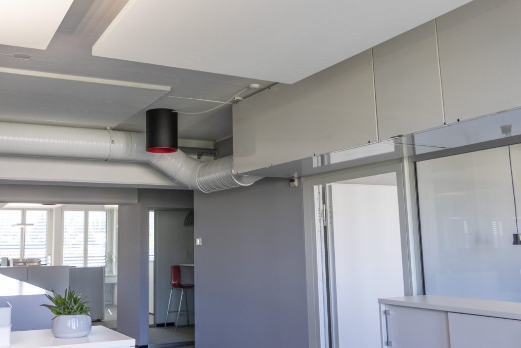 Bürointerieur mit grauen Wänden, offenem Arbeitsbereich, Belüftungssystem und einem schwarzen, zylindrischen Objekt an der Decke.