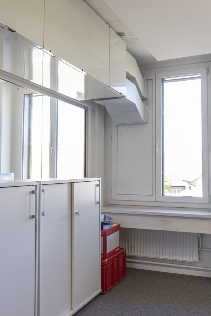 Weiße Schränke und ein helles Fenster in einem Eckbüro. Rote Aufbewahrungsboxen auf dem Boden.