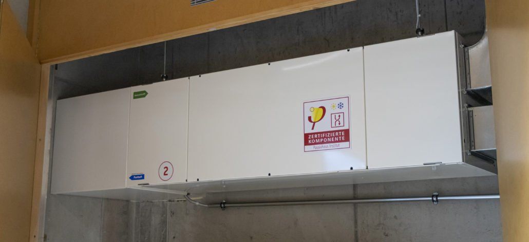 Weißes, rechteckiges Lüftungsgerät mit Beschriftung, montiert unter einem Holzbalken.
