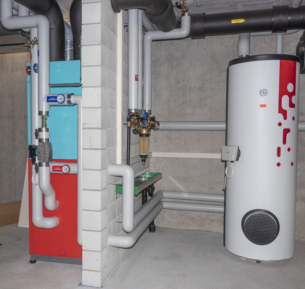 Heizsystem mit rotem und blauem Heizkessel, weißem Warmwasserbereiter und Verbindungsrohren in einem Betonraum.