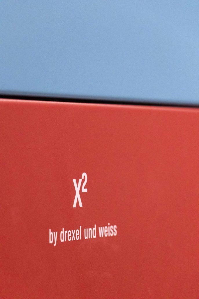 Rotes Kästchen mit weißem Schriftzug „X²“ und „by drexel und weiss“ auf blauem Hintergrund.