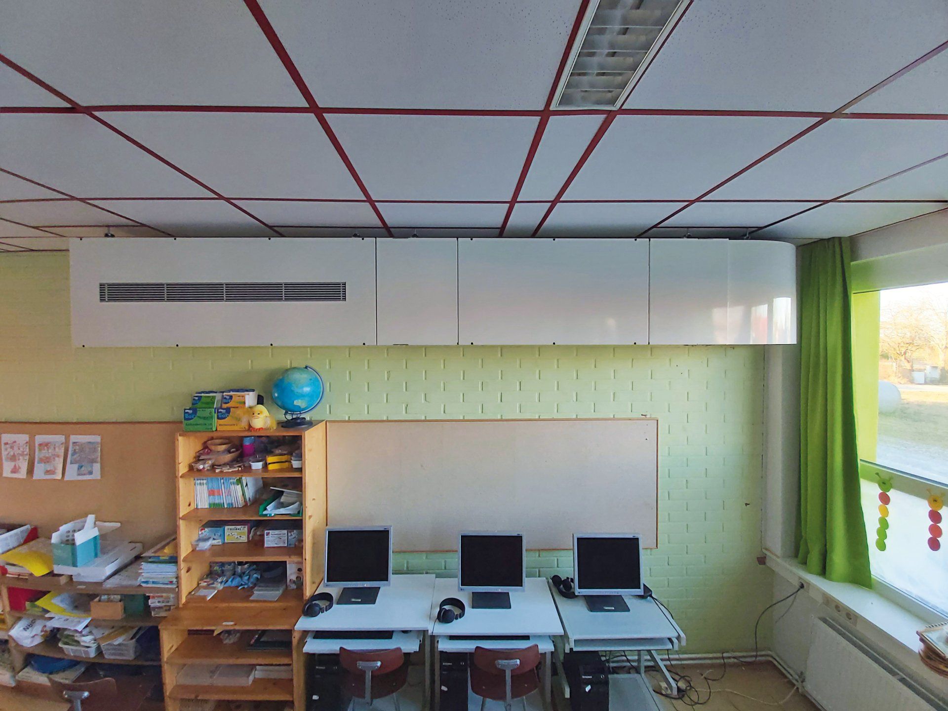 Klassenzimmer mit Computern, Whiteboard und Deckenlüftung. Holzregale, ein Fenster mit grünen Vorhängen.