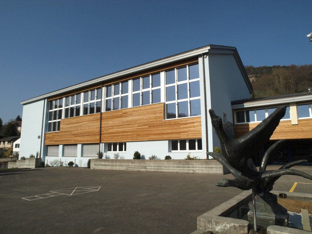 Schulgebäude mit hellblauen Wänden, Holzelementen, großen Fenstern und einer Vogelskulptur.