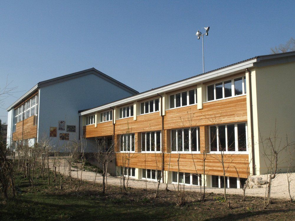Schulgebäude mit hellbrauner Holzverkleidung, blauen und gelben Wänden und mehreren Fenstern.