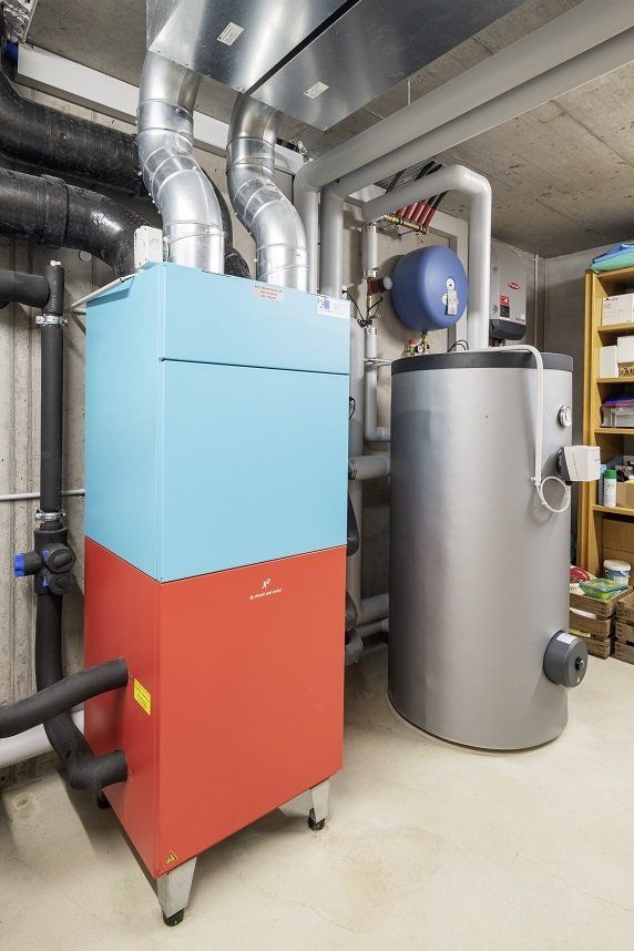 Blaue und rote Heizungsanlage mit silbernen Rohren und einem Grauwassertank in einem Hauswirtschaftsraum.