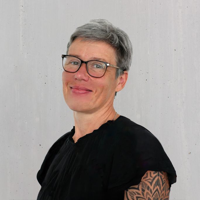 Person mit kurzen grauen Haaren und Brille lächelt, trägt ein schwarzes Oberteil; gemustertes Tattoo auf dem Arm.