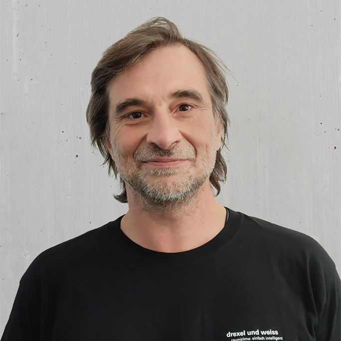 Mann mit braunen Haaren und Bart lächelt, er trägt ein schwarzes Hemd. Graue Wand im Hintergrund.