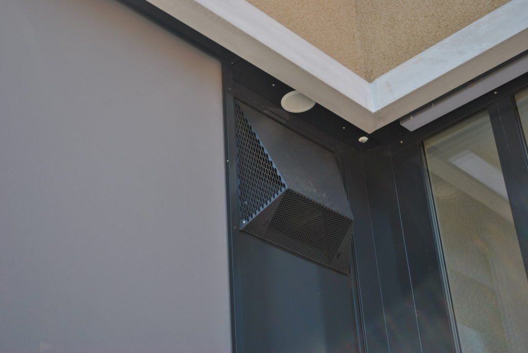 Ein metallenes Lüftungsgitter mit Wabenmuster an einer dunklen Gebäudeecke, in der Nähe von Decke und Fenster.