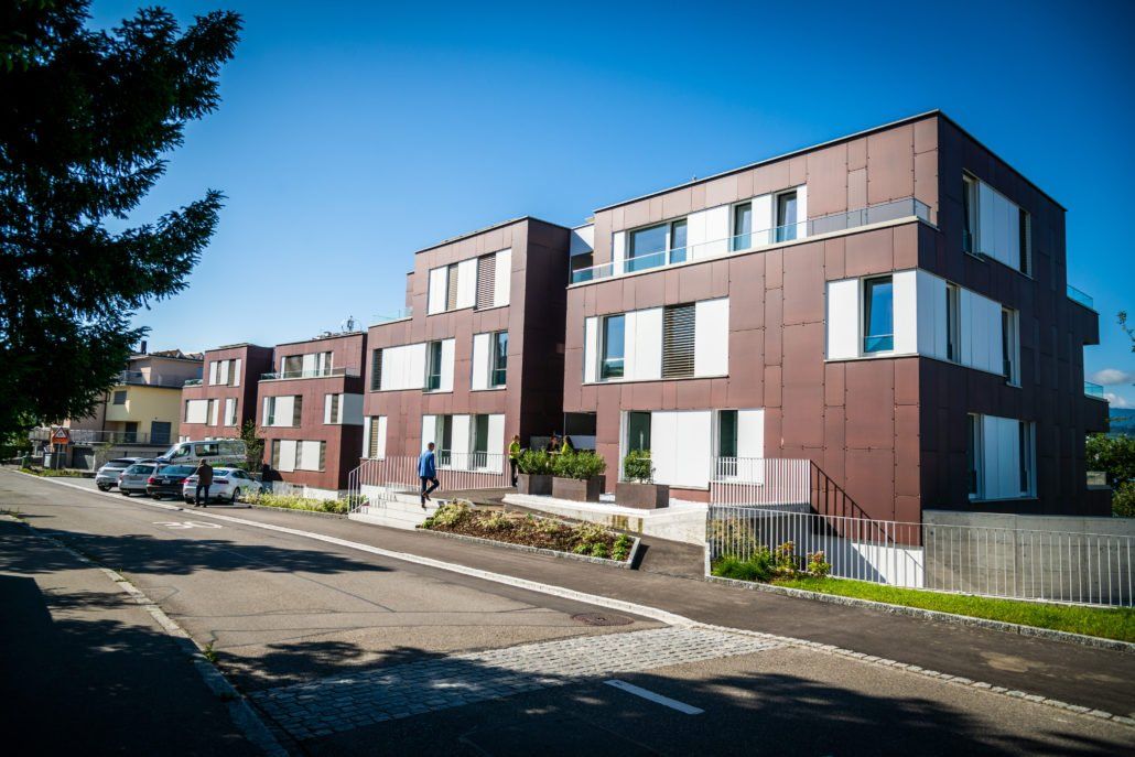 Braunes Wohnhaus mit weißen Fensterläden an einer von Bäumen gesäumten Straße unter blauem Himmel.