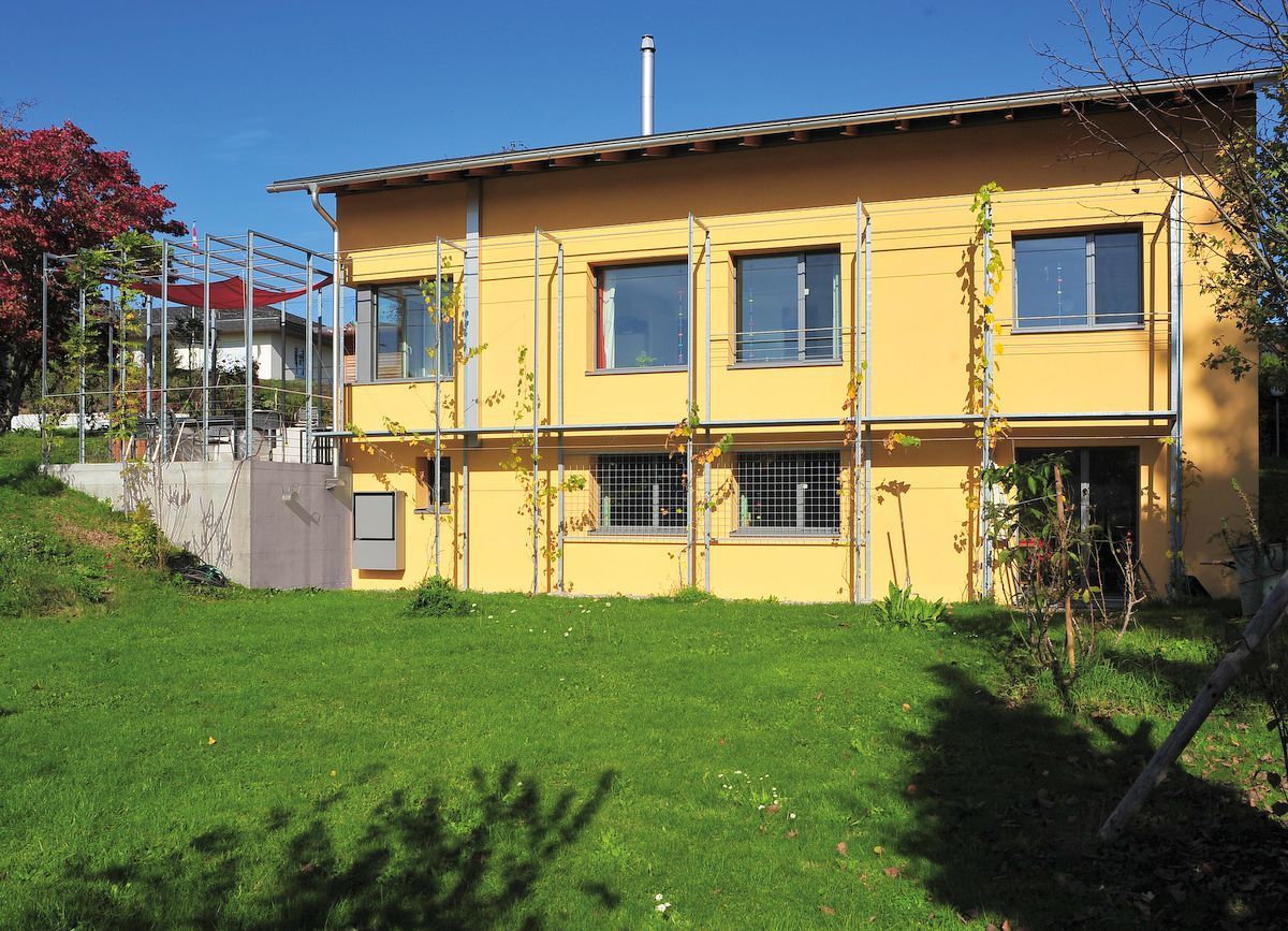Gelbes zweistöckiges Haus mit grünem Rasen im Garten. Weinreben ranken an Metallstützen empor. Strahlend sonniger Tag.