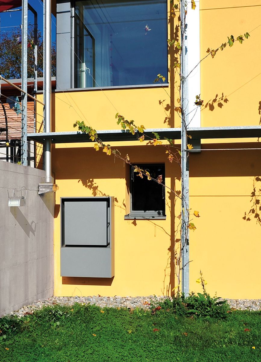 Gelbes Gebäude mit einem grauen Schrank, einem kleinen dunklen Fenster und einer Kletterpflanze.