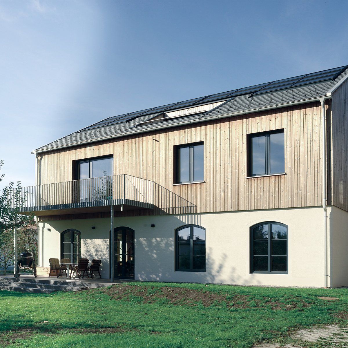Zweistöckiges Haus mit Holzverkleidung, Balkon und Bogenfenstern auf einem Rasen unter blauem Himmel.