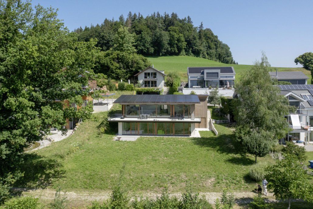 Modernes Haus mit Glaswänden und Solarpaneelen am Hang mit Blick auf einen Rasenhof und die umliegenden Bäume.