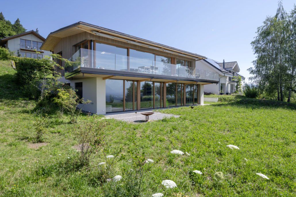 Modernes Haus mit großen Glasfenstern und Holzrahmen, mit Blick auf einen grünen Hügel.