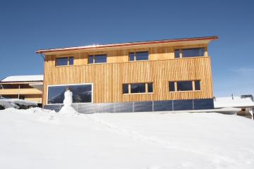 Holzhaus mit Solarpaneelen auf schneebedecktem Boden unter klarem, blauem Himmel.