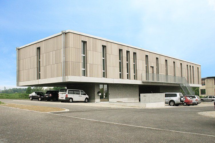 Modernes, rechteckiges Gebäude mit Holzfassade, das über einem Betonsockel auskragt. Autos parken darunter.