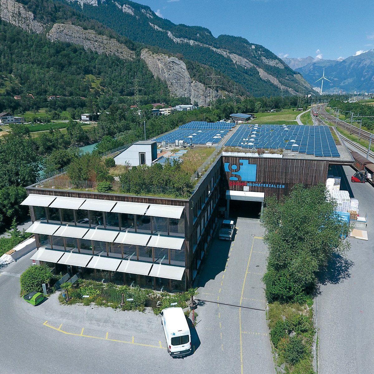 Gebäude mit Solaranlage, Gründach, Parkplatz, Berge im Hintergrund.