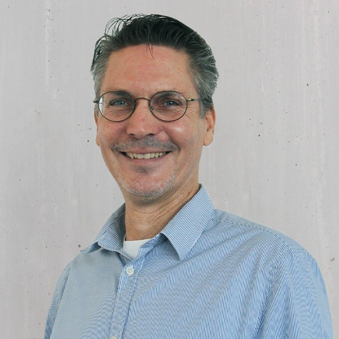 Ein Mann mit Brille lächelt in einem blau gestreiften Hemd vor einer weißen Wand.