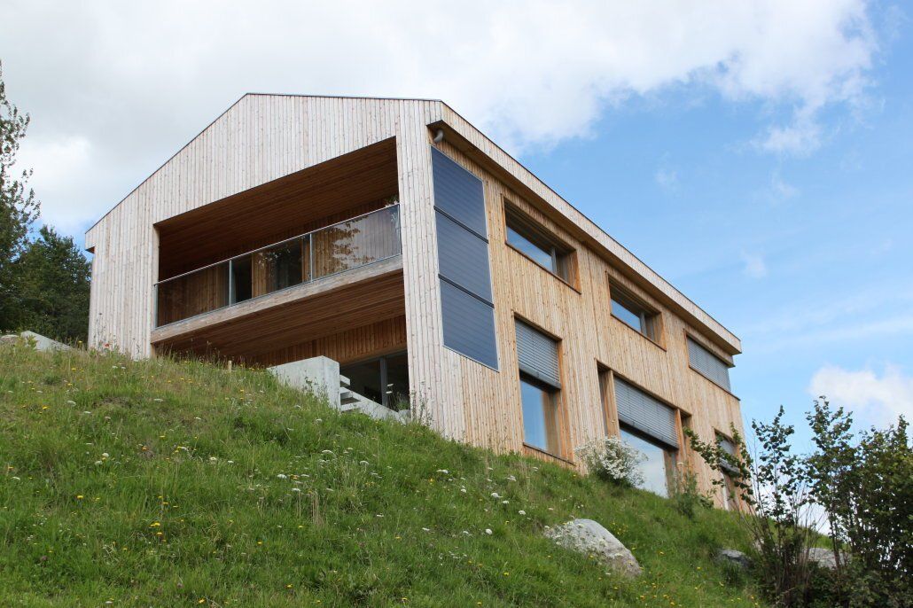 Modernes Holzhaus an einem grasbewachsenen Hang unter blauem Himmel. Balkon, große Fenster und Solarpaneele sind zu sehen.