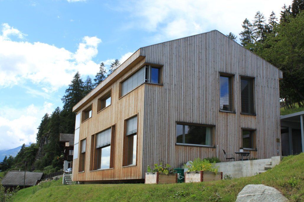 Ein Holzgebäude mit vielen Fenstern auf einem grasbewachsenen Hügel, umgeben von Bäumen unter blauem Himmel.