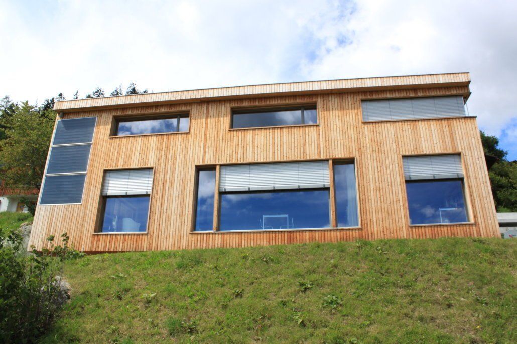 Modernes Holzhaus auf einem grasbewachsenen Hügel, mit großen Fenstern und Solarpaneelen.