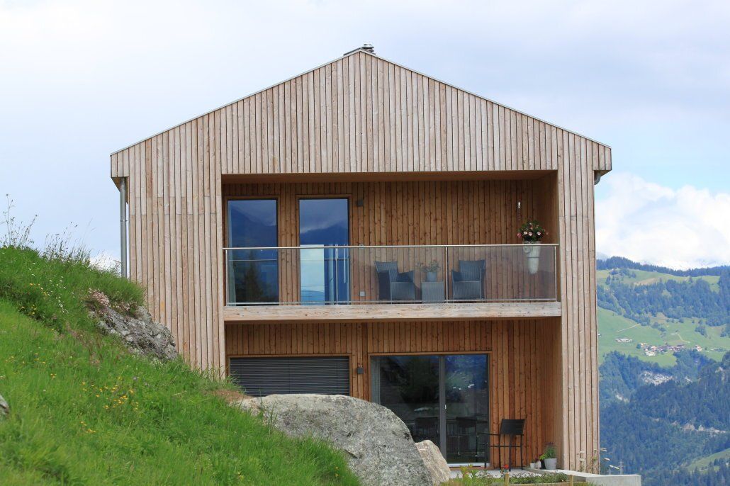 Zweistöckiges Holzhaus mit Balkon, erbaut an einem grasbewachsenen Hang. Bergkulisse.