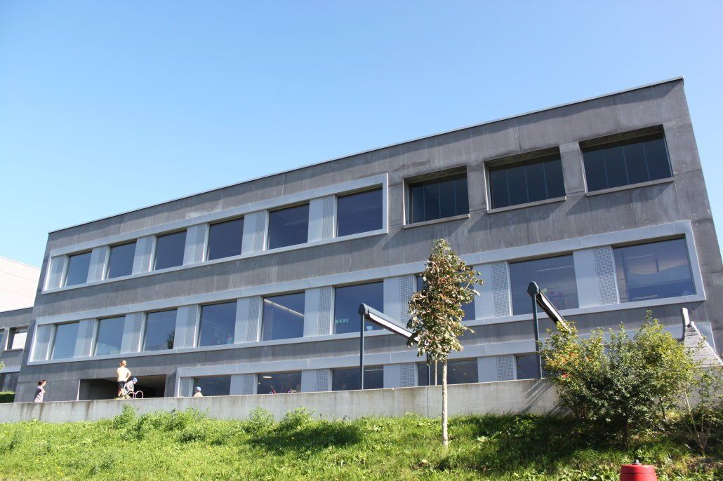 Graues dreistöckiges Gebäude mit vielen Fenstern unter blauem Himmel, im Vordergrund etwas Grün.