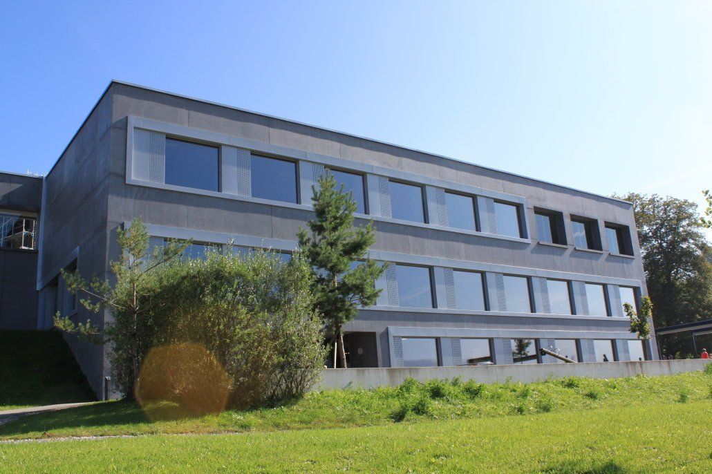 Graues Schulgebäude mit rechteckigen Fenstern auf einem grasbewachsenen Hügel unter blauem Himmel.