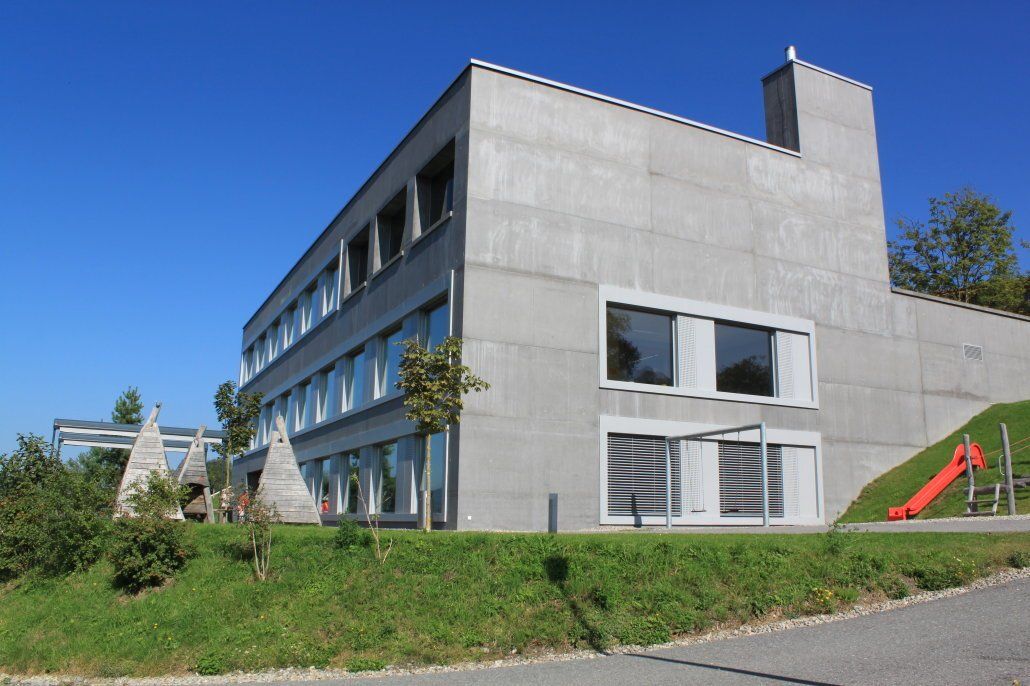 Modernes Schulgebäude aus Beton mit vielen Fenstern, grünem Rasen und blauem Himmel.
