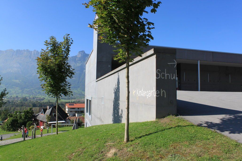 Außenansicht eines Gebäudes mit grauer Wand und grünem Rasen, mit Schildern, die auf „Schule“ und „Wort“ hinweisen.