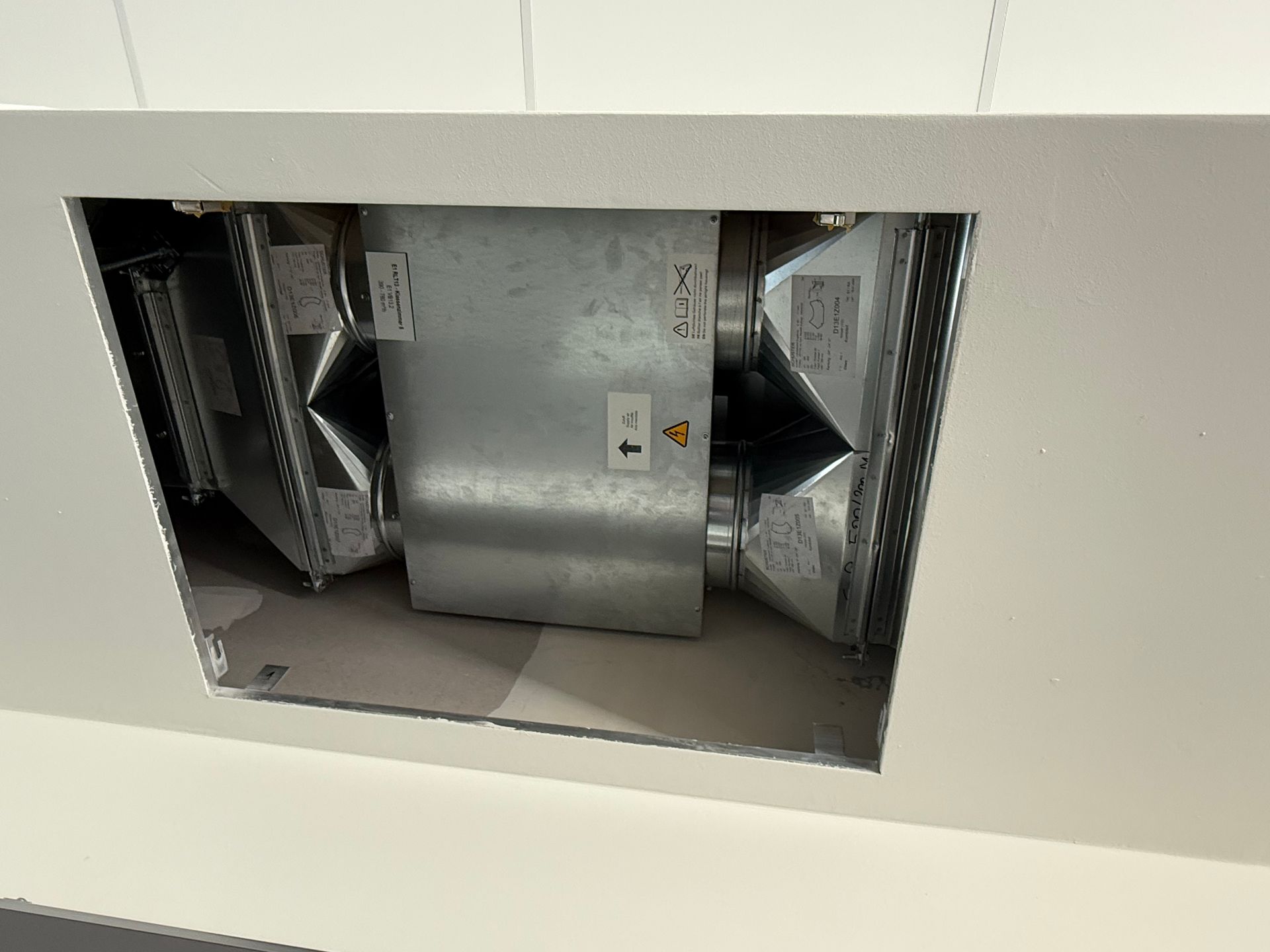 Ein freiliegendes Lüftungskanalsystem innerhalb einer weißen Decke, das eine rechteckige Öffnung und metallische Lüftungskanäle zeigt.