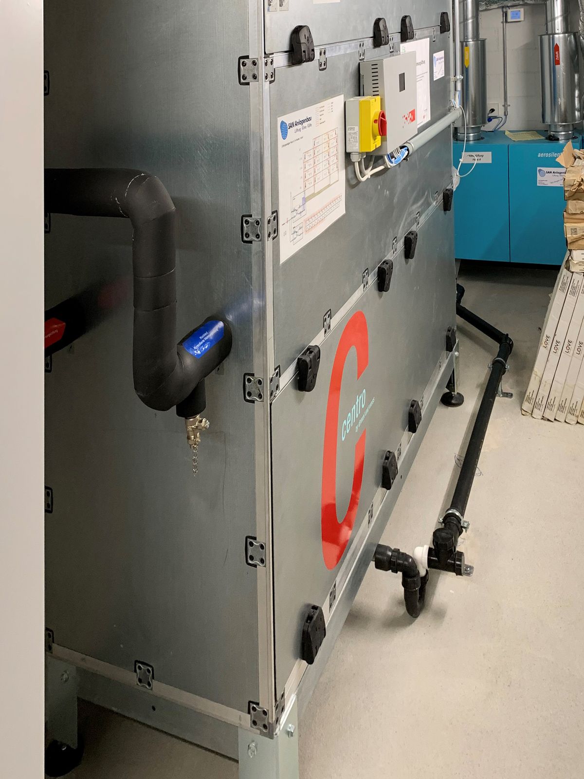 Metallene Klimaanlage mit schwarzen Rohrleitungen und rotem Logo in einer industriellen Umgebung.