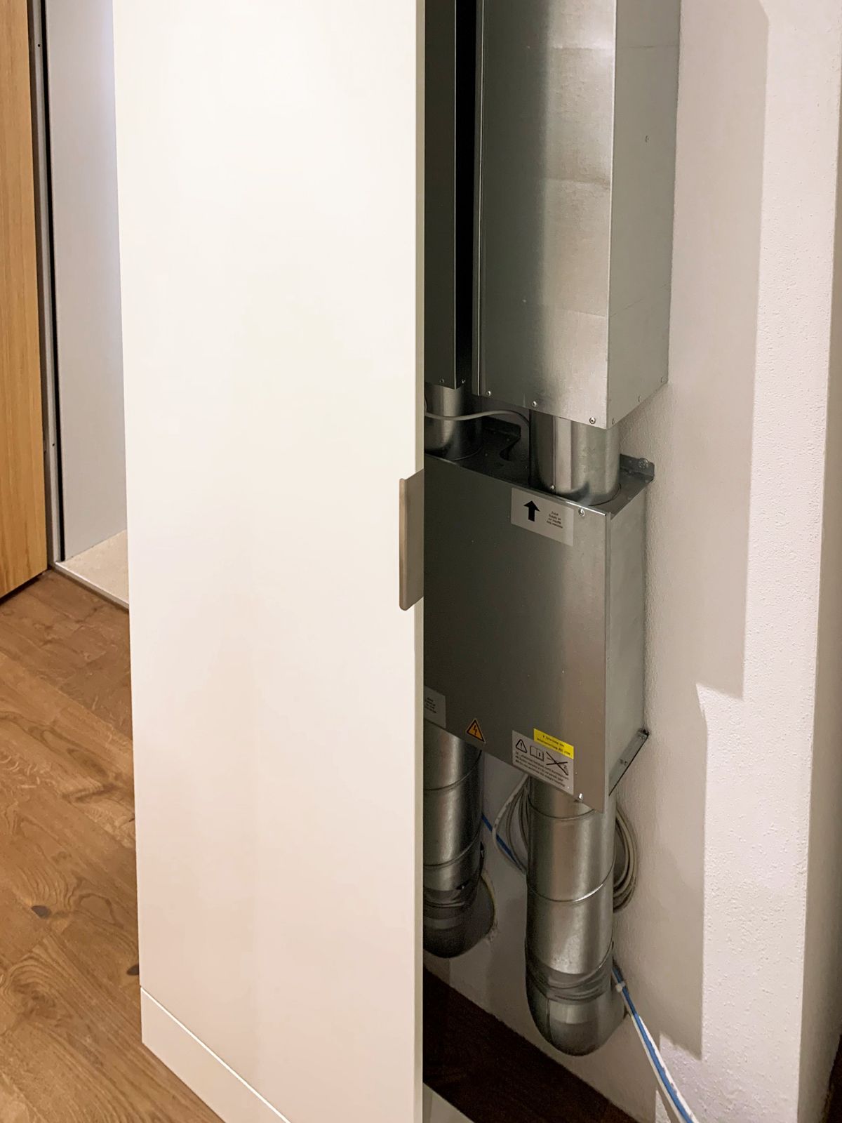 Weißer Schrank mit offener Tür, hinter der sich ein metallisches Lüftungssystem verbirgt, vor einer weißen Wand und einem Holzboden.