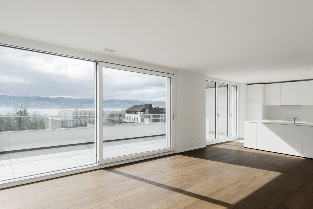 Großzügiges, modernes Apartment mit großen Fenstern, weißen Wänden und Holzböden mit Blick auf eine wolkige Landschaft.
