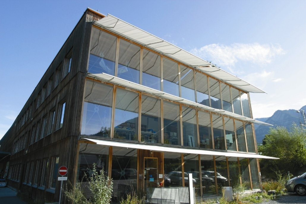 Modernes Gebäude mit großen Glasfenstern, Holzbalken und Solarpaneelen. Im Hintergrund Berge.
