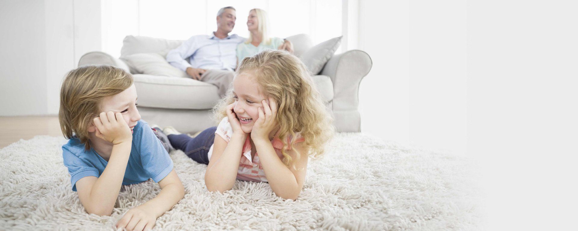 Zwei Kinder lachen auf einem weißen Teppich, die Großeltern auf dem Sofa. Helles, gemütliches Wohnzimmer.