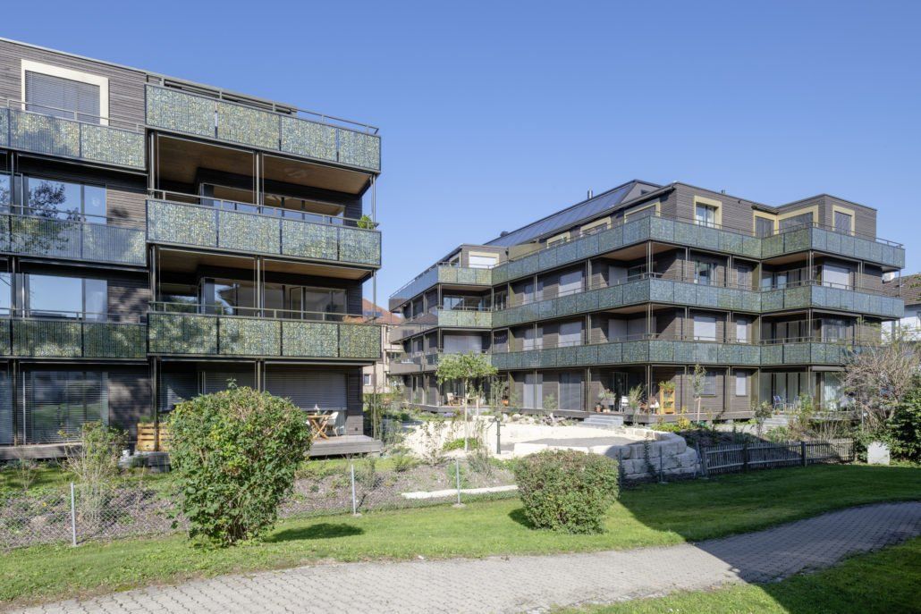 Zwei moderne Wohnhäuser mit Holzbalkonen, eingebettet in einen begrünten Innenhof an einem sonnigen Tag.