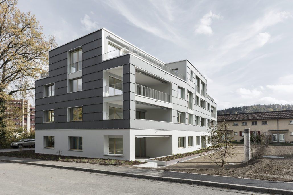 Modernes mehrstöckiges Gebäude mit grau-weißer Fassade, Balkonen und lichtdurchfluteten Innenräumen.