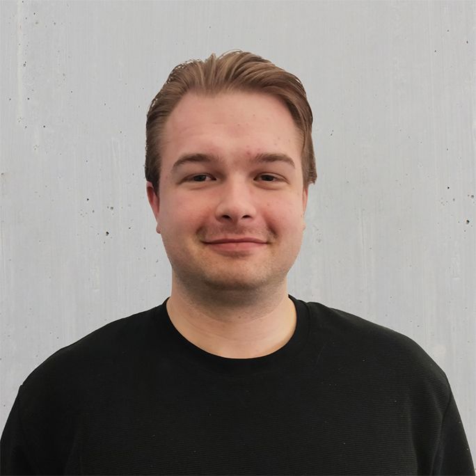 Ein Mann mit heller Haut und blonden Haaren in einem schwarzen Pullover lächelt vor einem grauen Hintergrund.