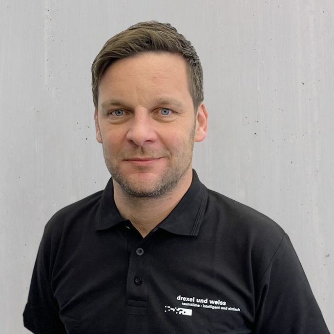 Ein Mann in einem schwarzen Poloshirt lächelt und posiert vor einer Betonmauer.