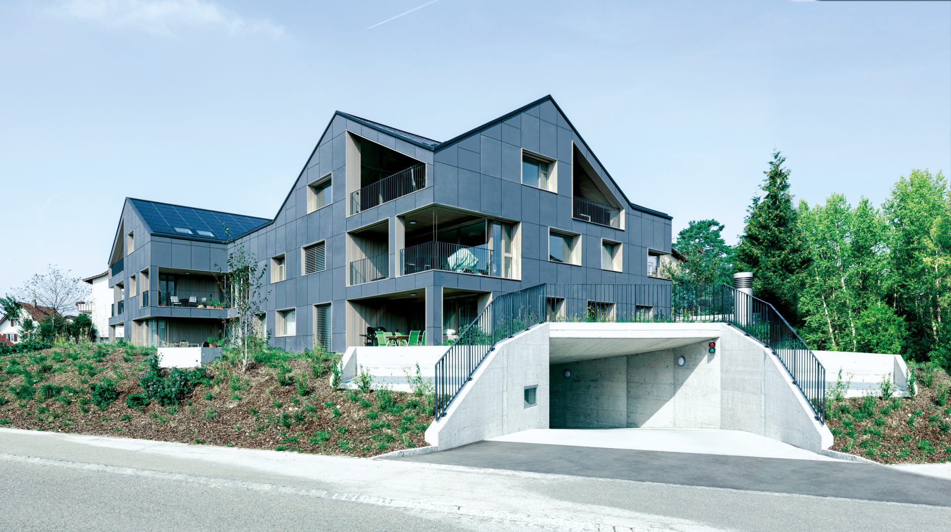 Dunkelgraues, modernes Apartmentgebäude mit schrägem Dach, Balkonen und einem gewölbten Eingang zum Parkplatz.
