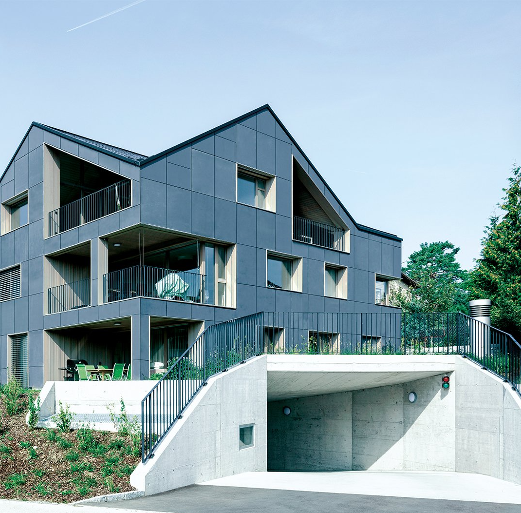 Modernes graues Mehrfamilienhaus mit Balkonen und überdachter Parkplatzeinfahrt.