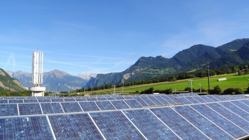 Solarpaneele auf einem Feld mit Windkraftanlage und Bergen unter blauem Himmel.