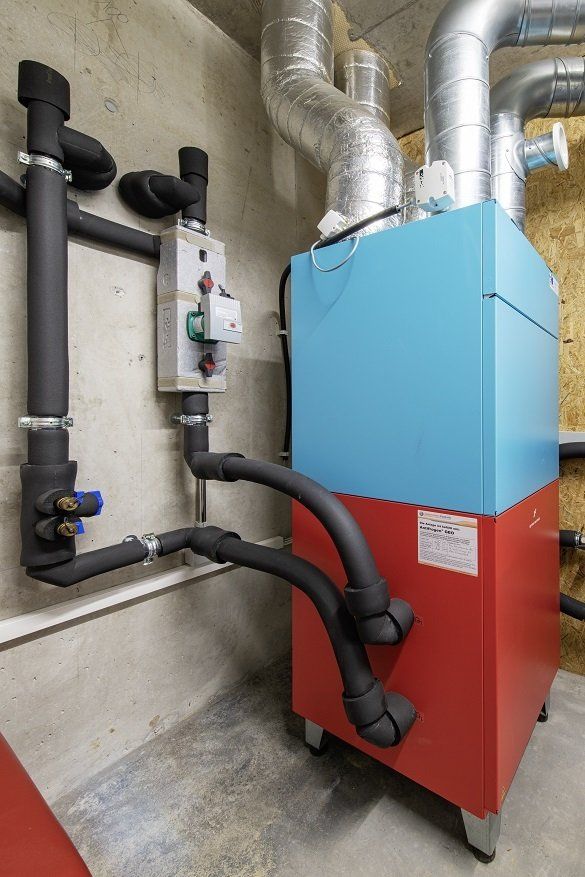 Industrielle Heizanlage in Blau und Rot mit schwarz isolierten Rohren und grauem Schaltkasten in einem Betonraum.