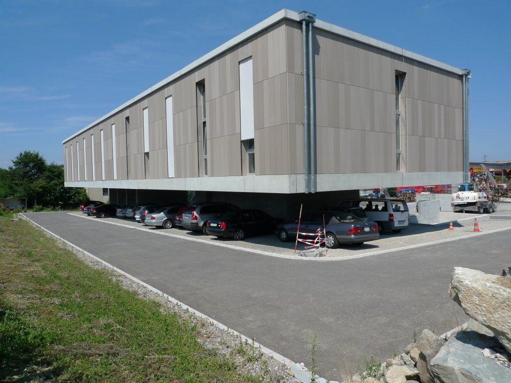 Ein rechteckiges Gebäude, das auf hydraulischen Hebern angehoben ist, darunter parken Autos. Graue Fassade, blauer Himmel.