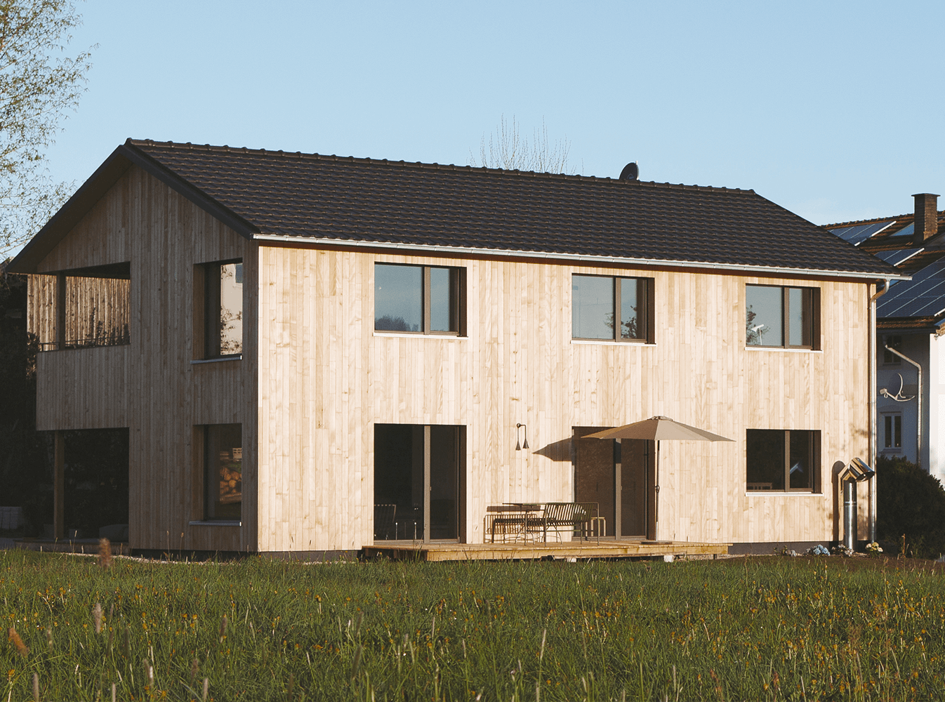 Zweistöckiges, holzverkleidetes Haus mit dunklem Dach und großen Fenstern, eingebettet in ein grasbewachsenes Feld.