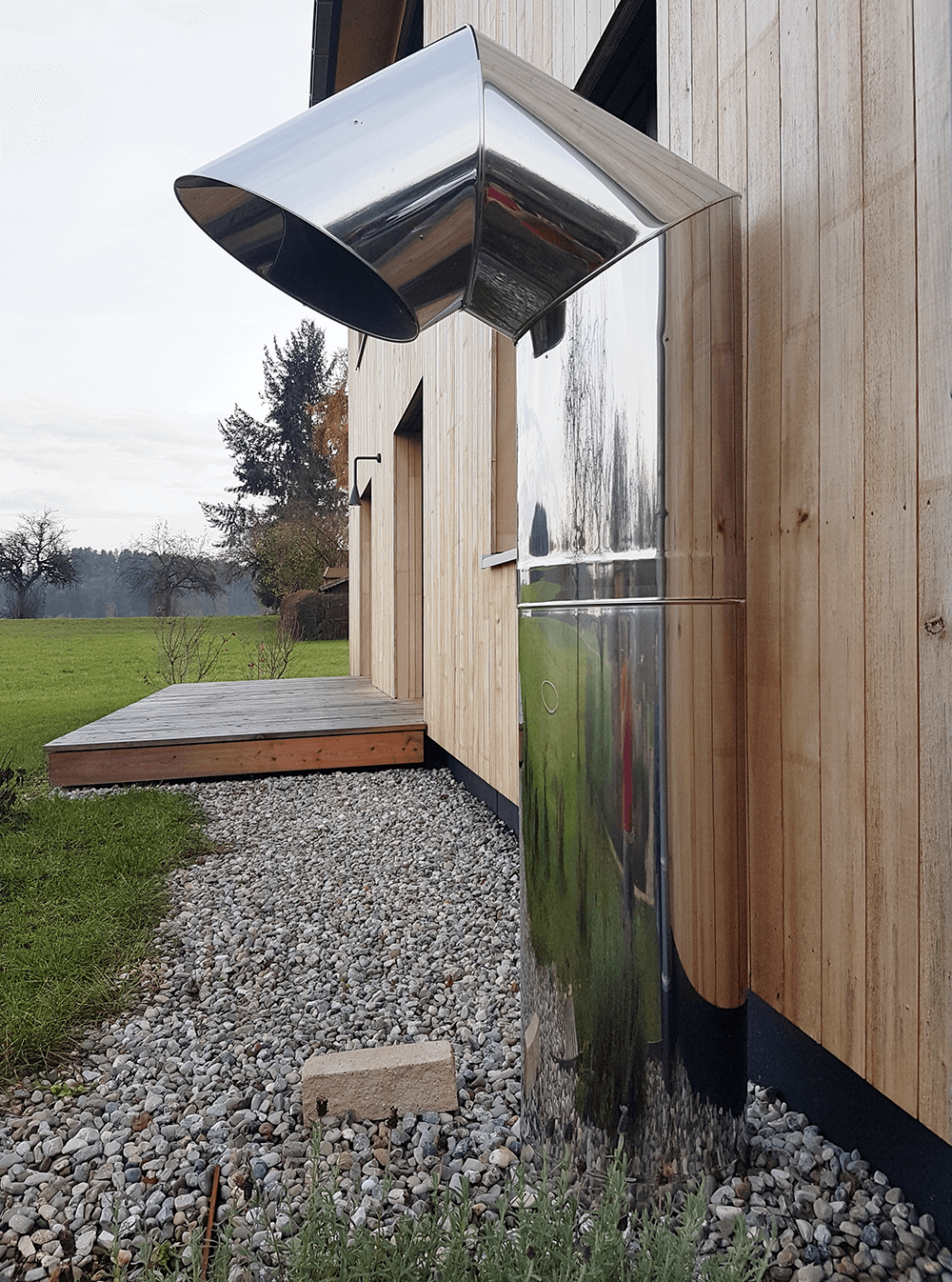 Ein glänzender Metallkamin an der Holzfassade eines Gebäudes, der sich über einen Kiesweg erstreckt, neben einer kleinen Holzveranda.